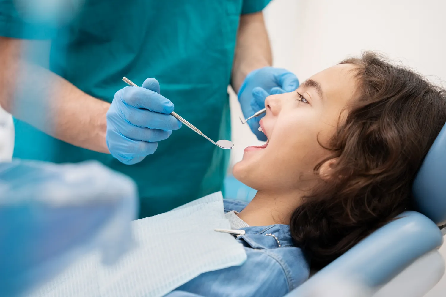 A dentist in teal scrubs and blue gloves holds a dental instrument near a patient's mouth, who is reclining in a dental chair.